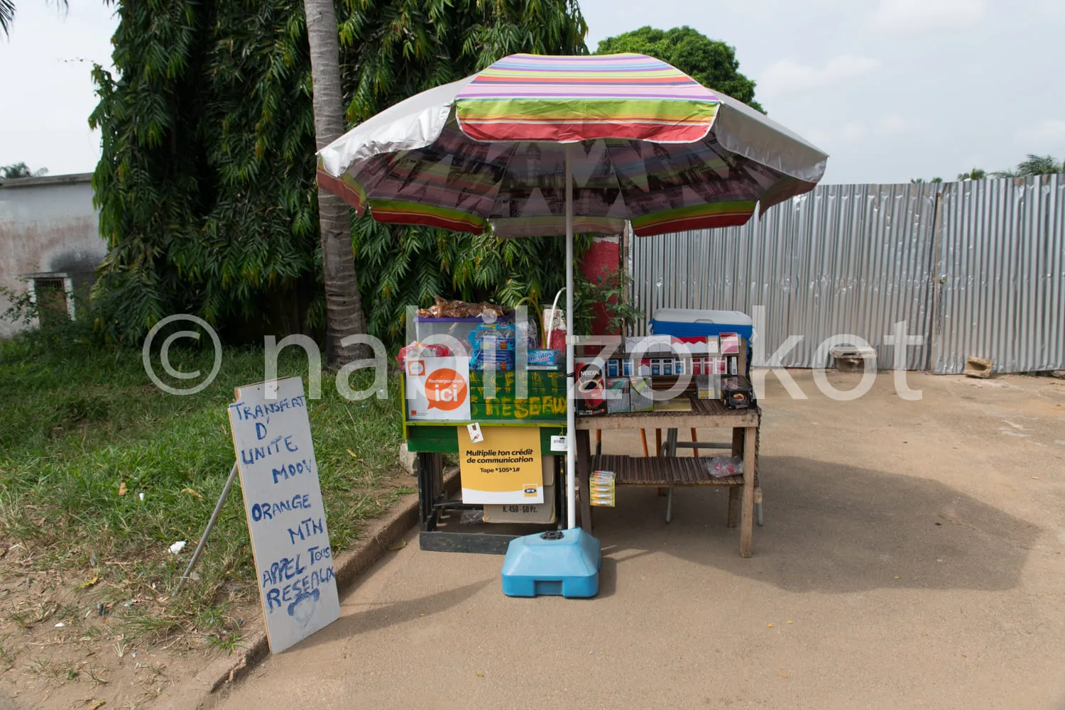 Africa photo — Un petit stand de recharge de téléphone mobile protégé par un parasol coloré, avec une affiche indiquant les services disponibles comme
