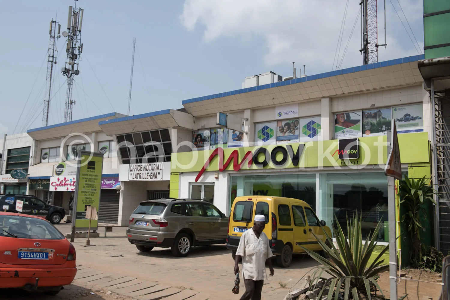 Africa photo — Image montrant un bâtiment MédiMarket avec des enseignes commerciales colorées, des voitures garées devant et un homme marchant sur un