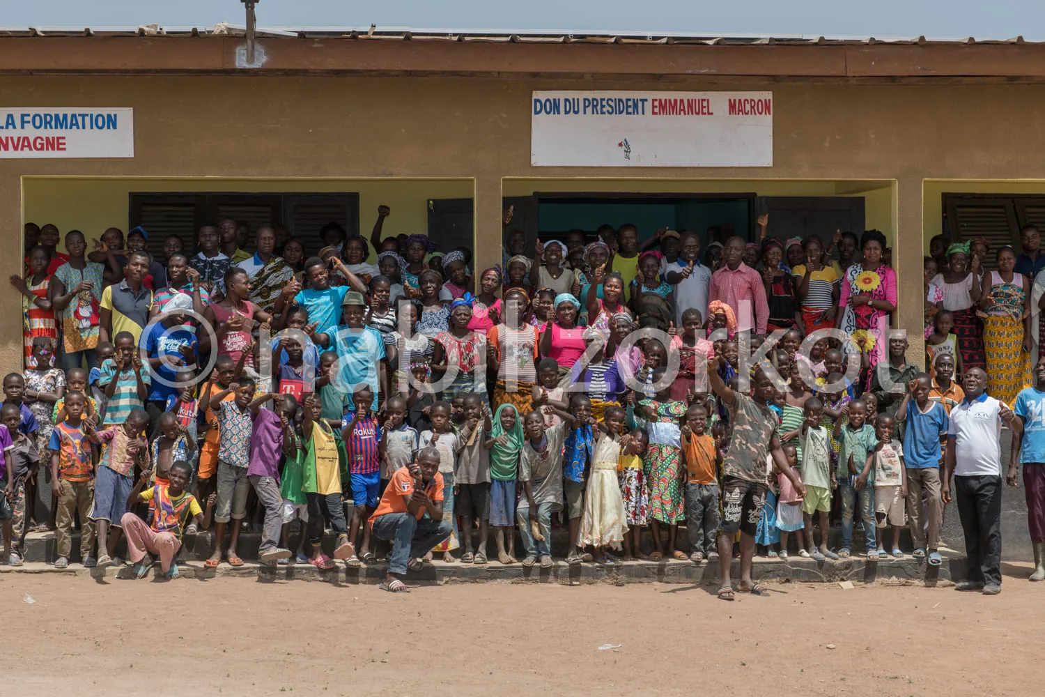Africa photo — Un groupe joyeux composé d’enfants et d’adultes posant devant un bâtiment jaune avec une bannière indiquant 'Don du Président Emmanuel