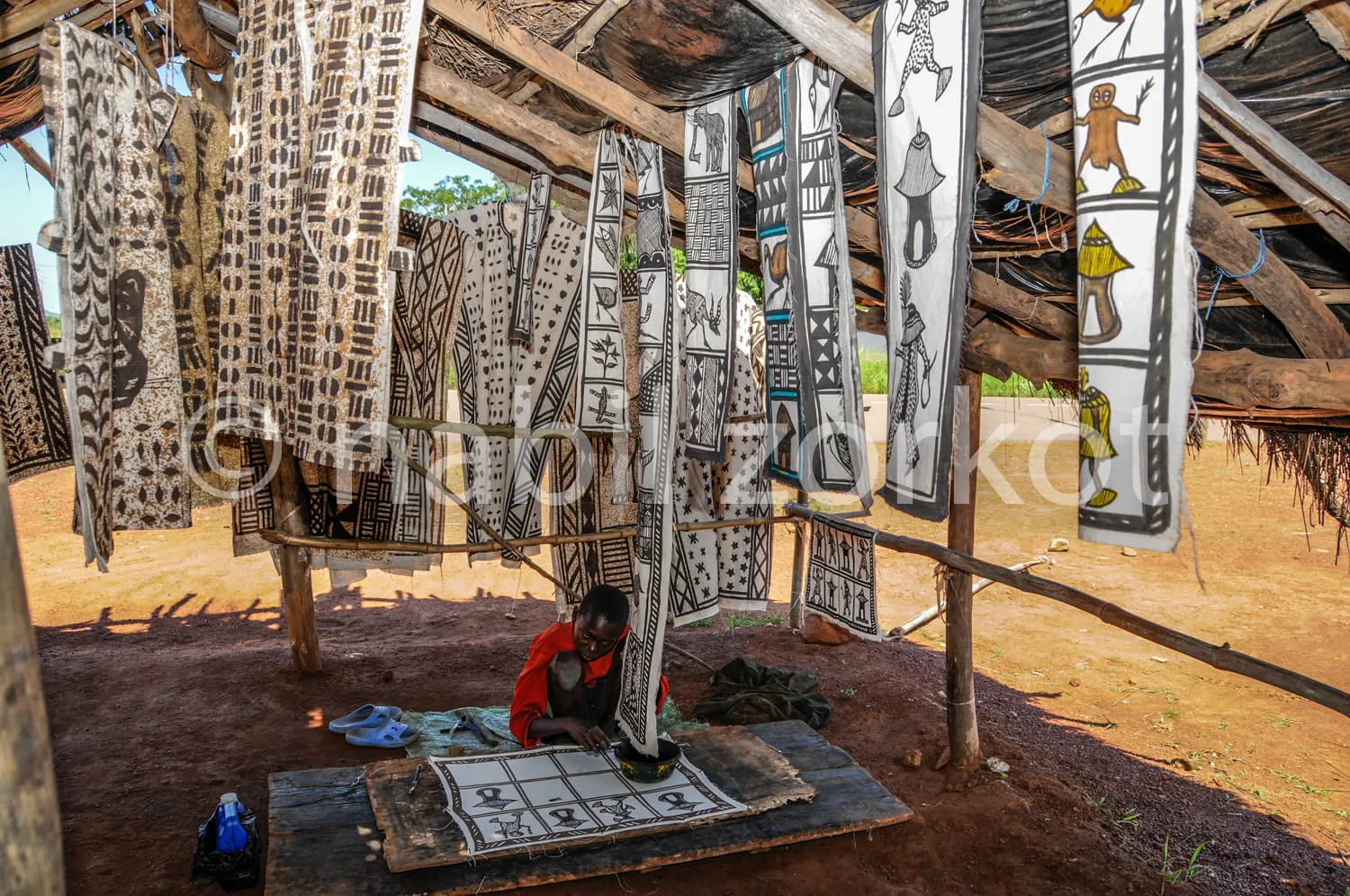 Africa photo — Un artisan assis sous une structure en bois ouverte, travaillant sur un tissu étendu sur une table, entouré de nombreux tissus colorés