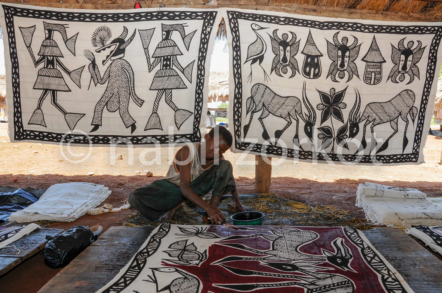 Africa photo — Un artisan est assis par terre dans un atelier, en train de travailler sur un tissu coloré aux motifs traditionnels. Deux autres tissus