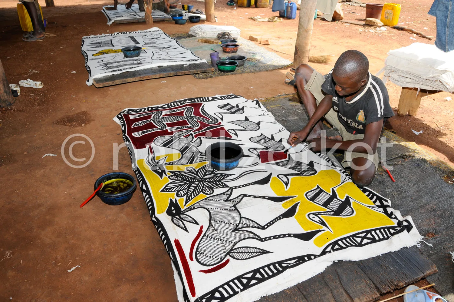 Africa photo — Un artisan est accroupi sur une surface en bois, en train de broder minutieusement un grand tissu coloré aux motifs géométriques et sym