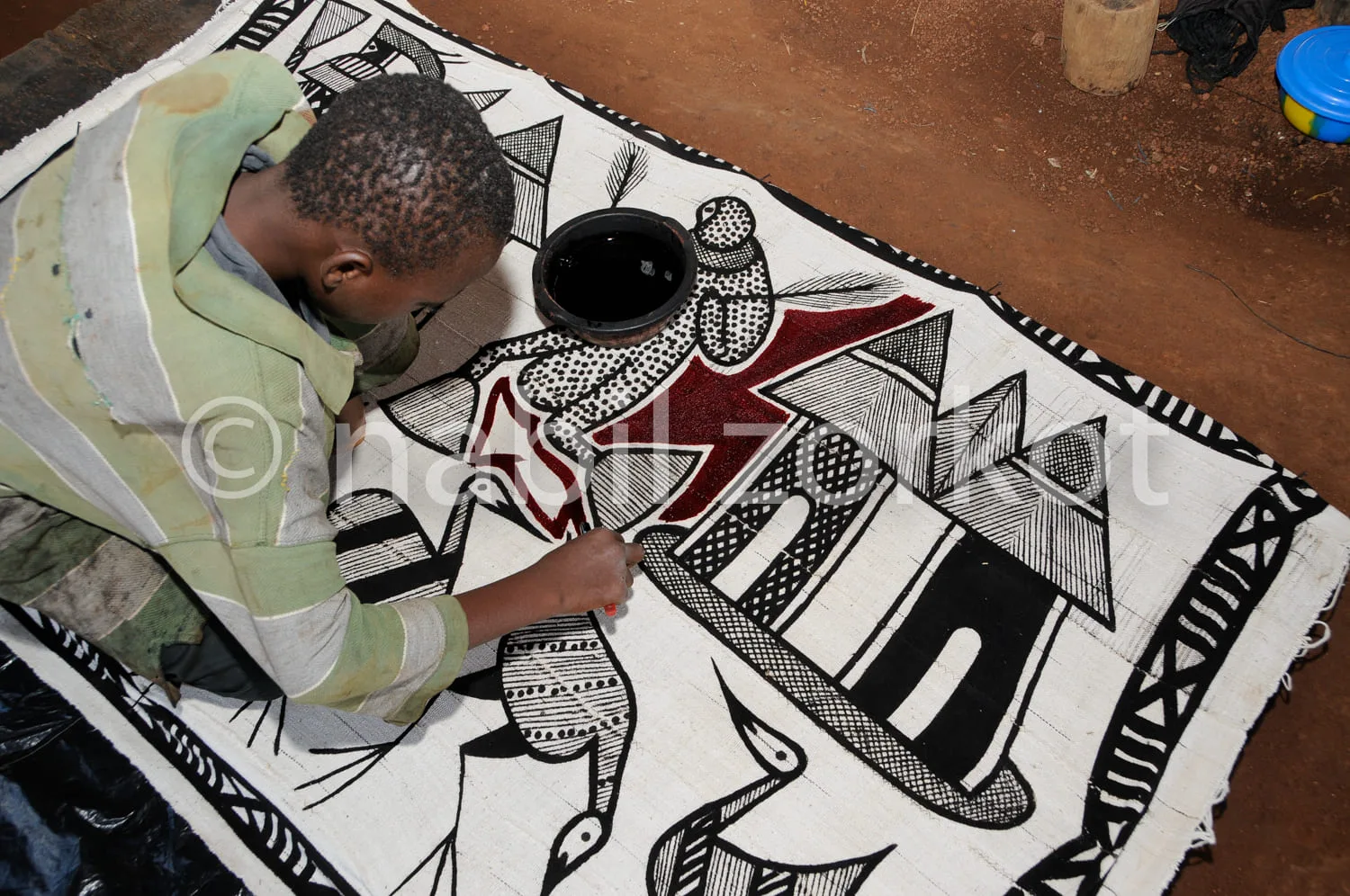 Africa photo — Un artisan est penché sur un grand tissu blanc et coloré, brodant minutieusement des motifs géométriques et des symboles traditionnels
