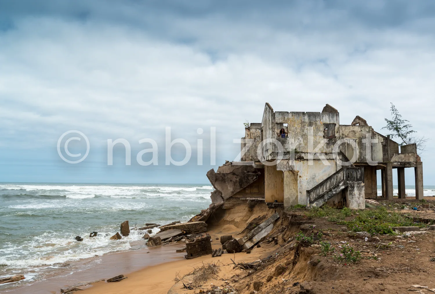 Africa photo — Un bâtiment partiellement effondré se dresse sur une plage, avec des vagues qui viennent lécher ses fondations. Une personne se tient à