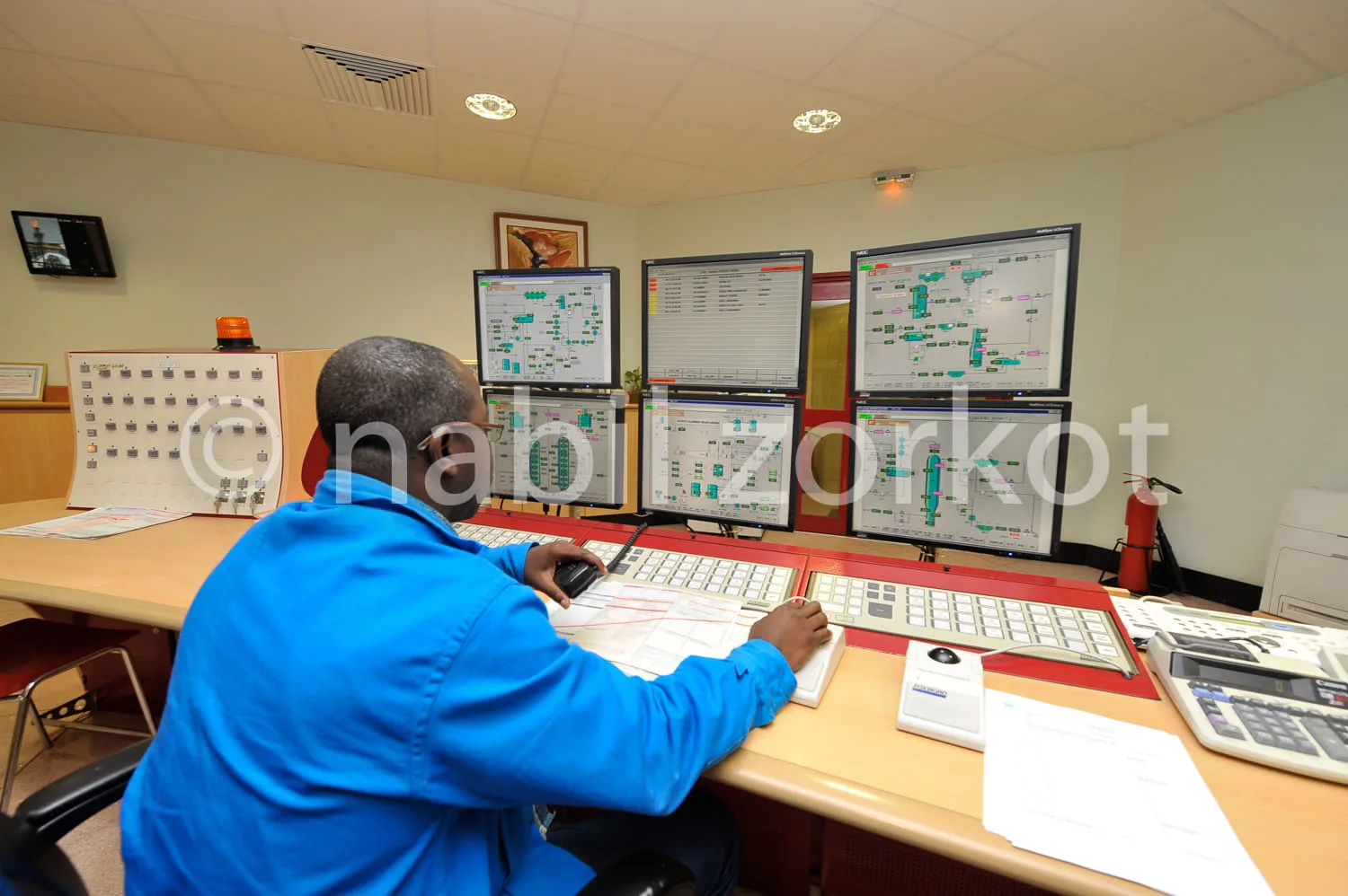 Africa photo — Un technicien en bleu, dos tourné, travaille devant trois écrans affichant des schémas électriques et des données techniques, avec des