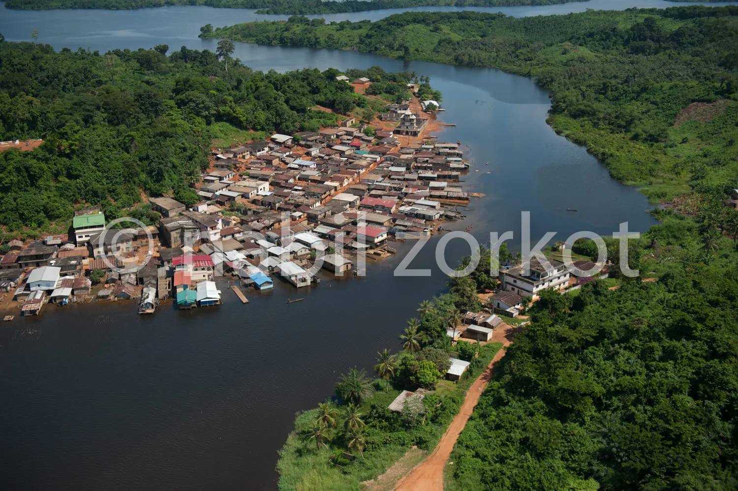 Africa photo — Vue aérienne d’un village riverain entouré de végétation luxuriante, avec des maisons colorées alignées le long d’une rivière sinueuse,