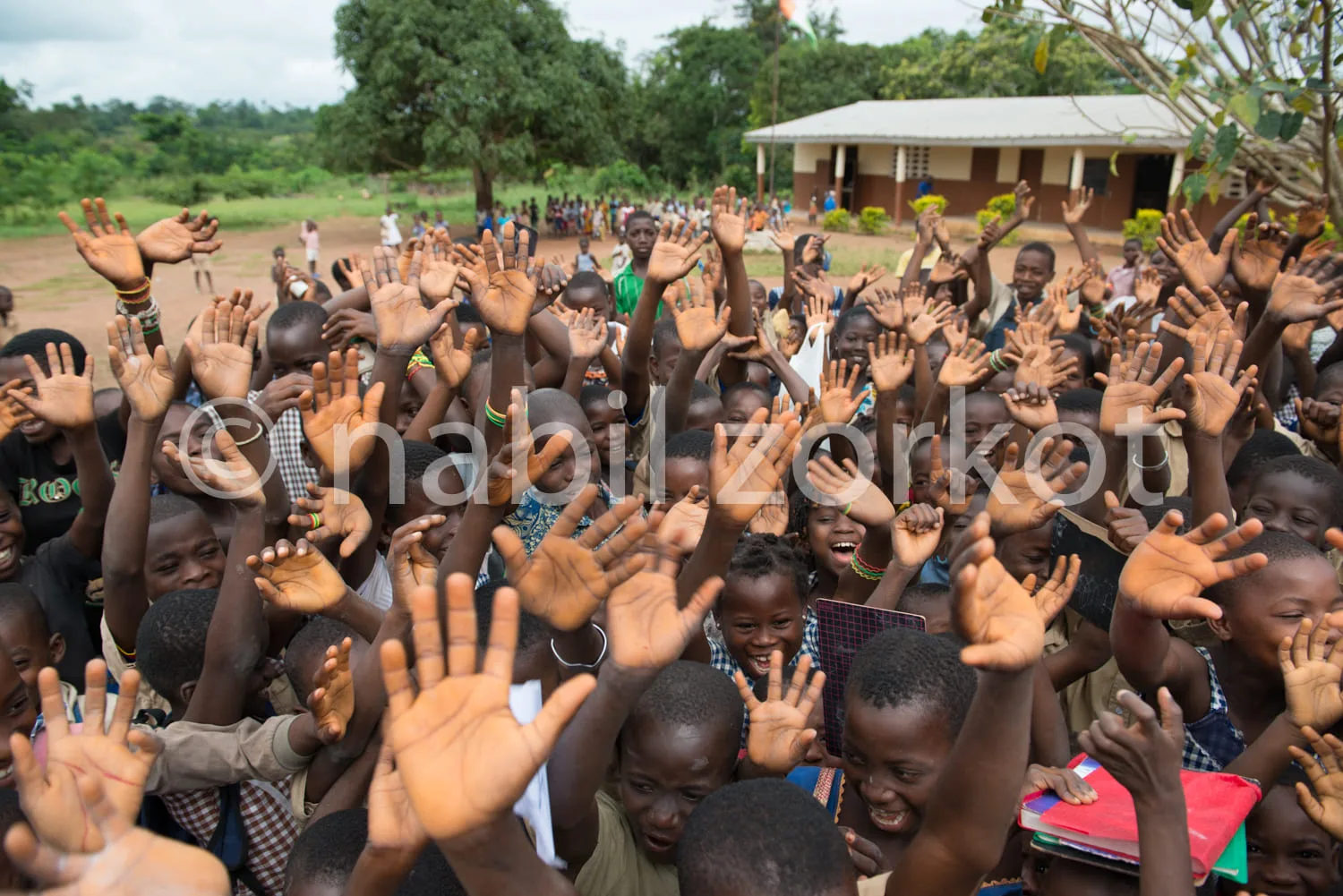 Africa photo — Un groupe d'enfants et d'adultes, souriants et enthousiastes, lèvent les mains en signe de joie devant une école en milieu rural, sous