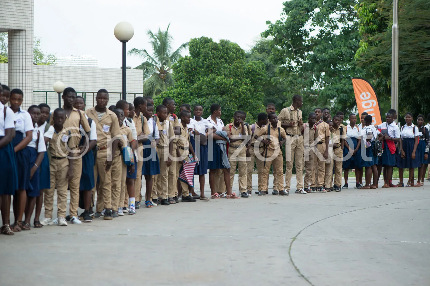 Africa photo — Groupe d’élèves en uniforme scolaire, debout en rang serré sur une allée en béton, avec un adulte en tenue similaire au centre. À droit