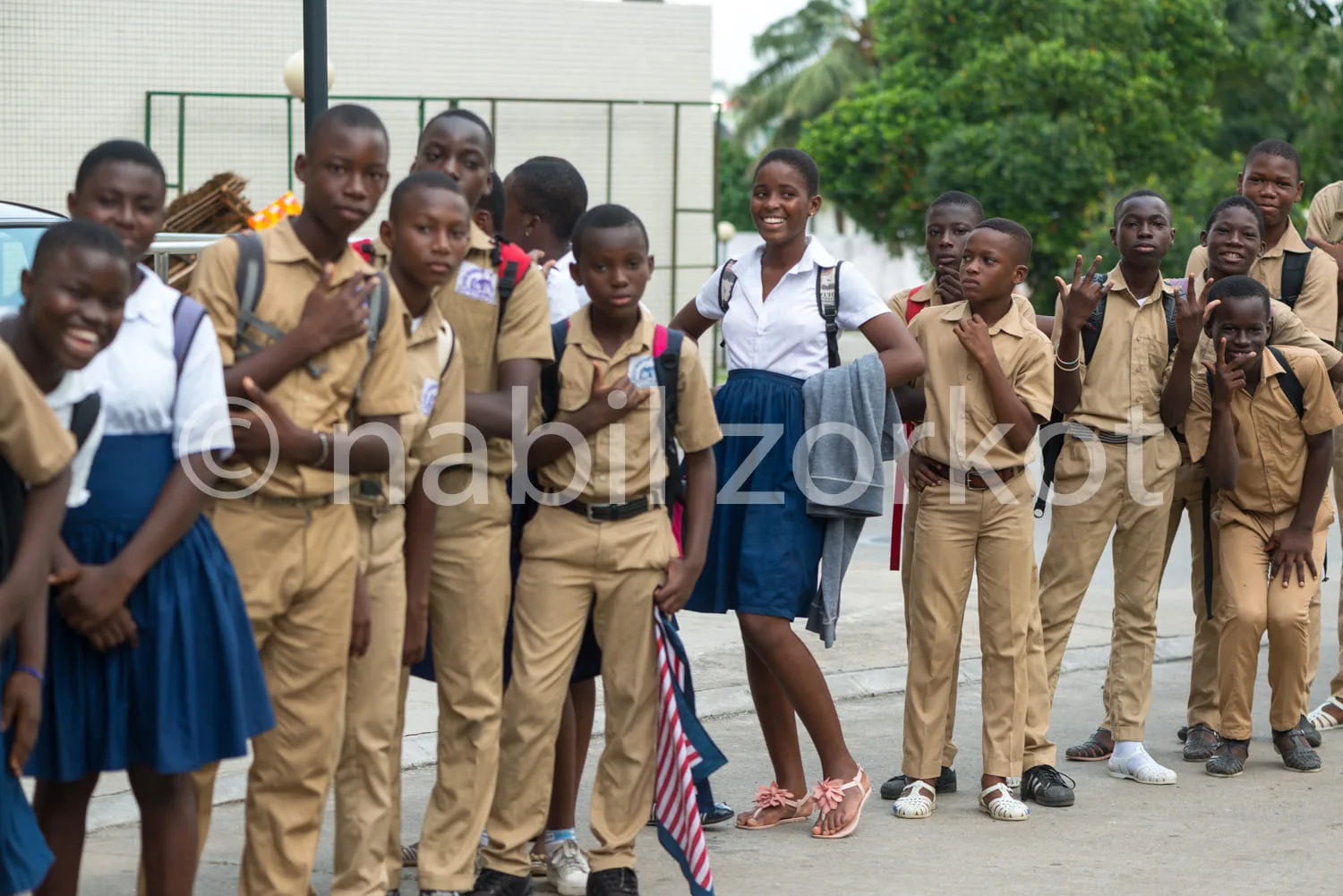 Africa photo — Un groupe d’enfants souriants, vêtus d’uniformes scolaires beiges et bleus, posent devant un bâtiment institutionnel. Certains tiennent