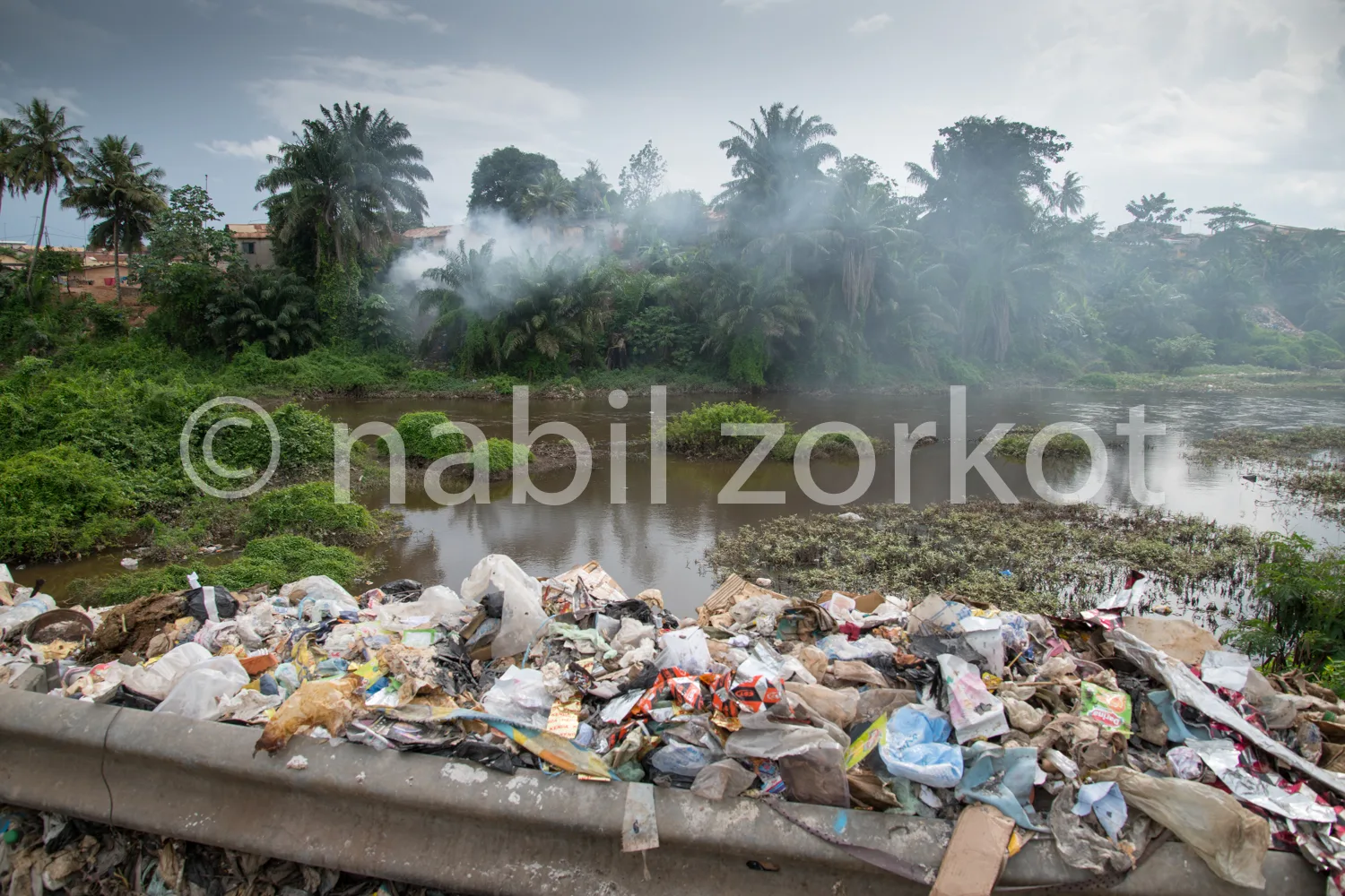 Africa photo — Image montrant une grande quantité de déchets plastiques et divers matériaux jetés sur un pont en béton, avec une rivière polluée et de