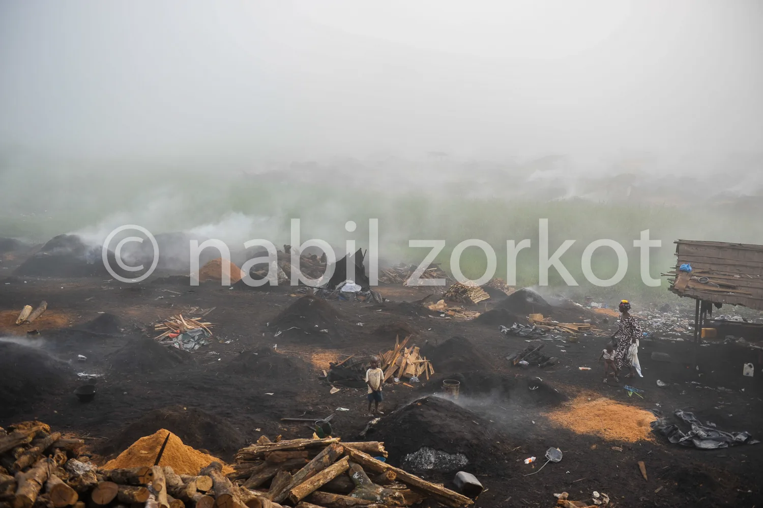 Africa photo — Un homme manipule des outils près de tas de charbon en combustion, entouré de bois carbonisé et de fumée épaisse, dans un environnement