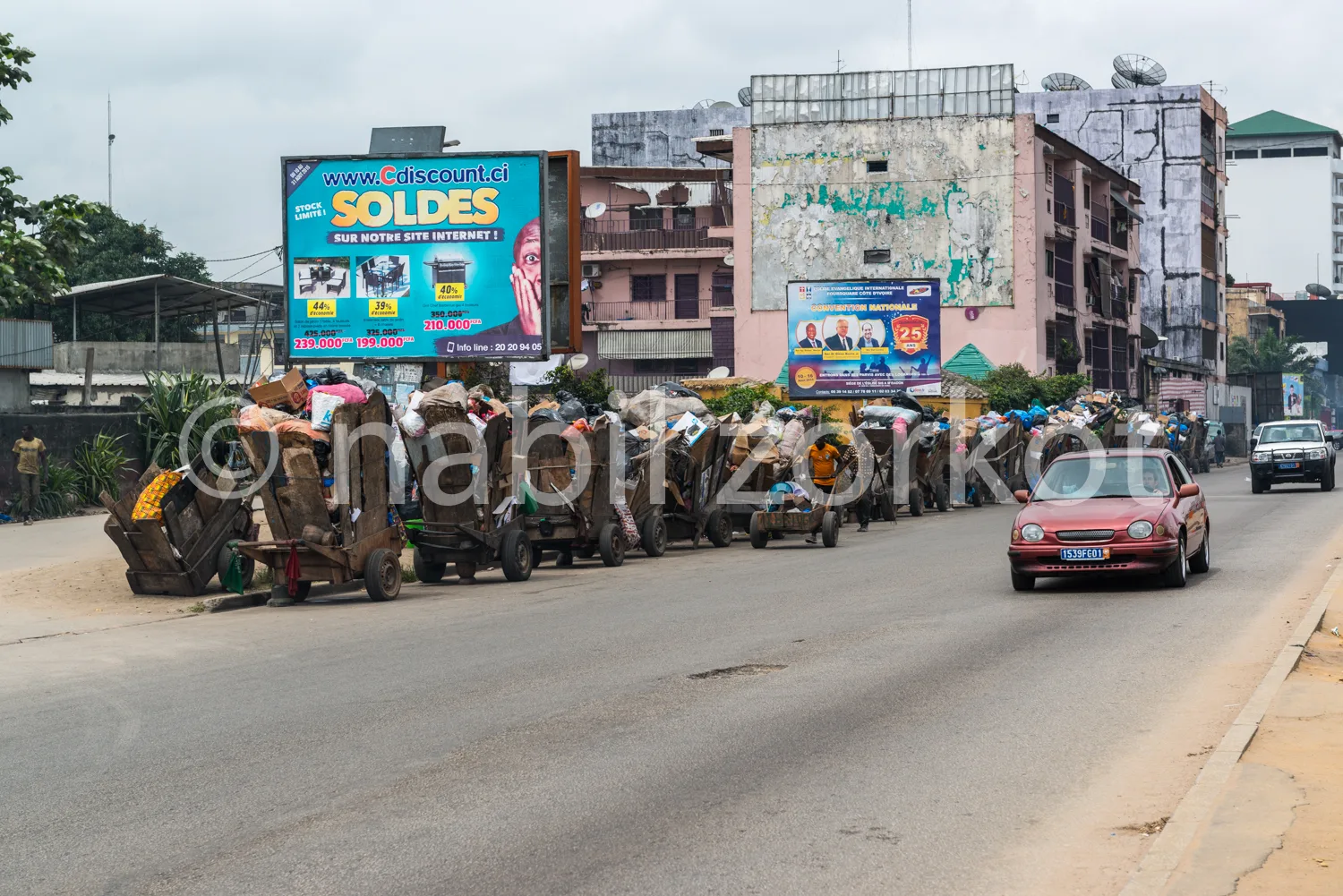 Africa photo — Une rue animée où plusieurs charrettes remplies de sacs et de marchandises avancent lentement au milieu d’un trafic léger. En arrière-p