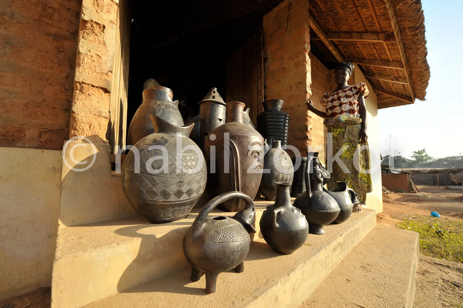 Africa photo — Une femme vêtue d’une robe colorée et imprimée se tient près d’une maison en terre, à côté d’une série de poteries artisanales de diffé
