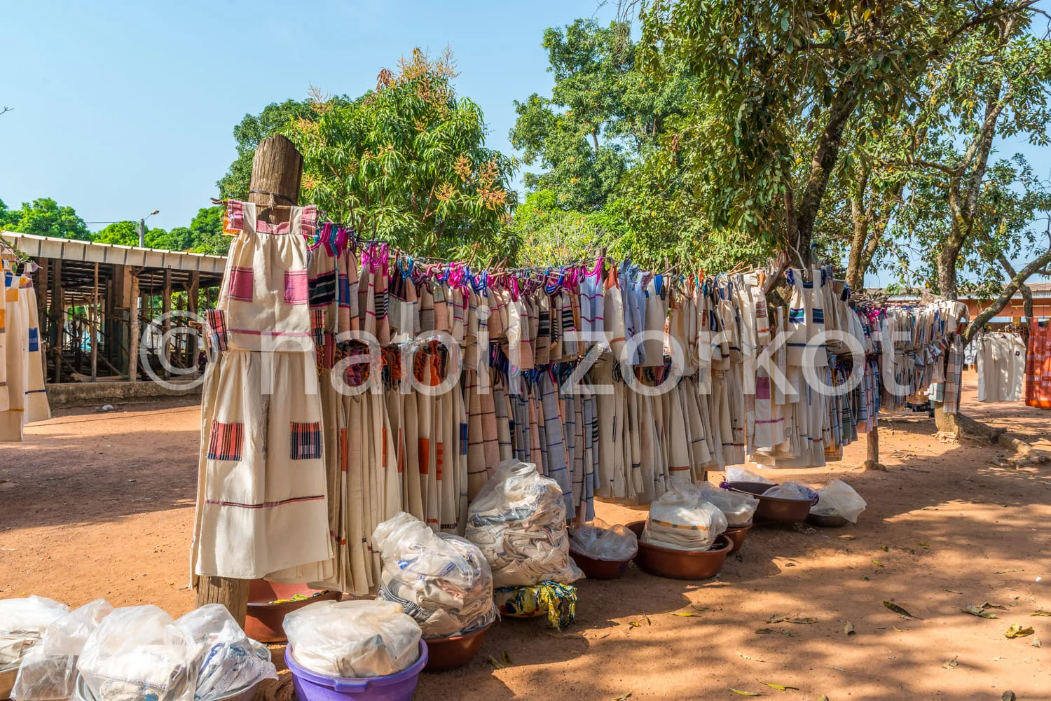 Africa photo — Une rangée de tissus colorés et imprimés suspendus à des perches en bois, avec des sacs et paniers remplis de vêtements au sol, sous de