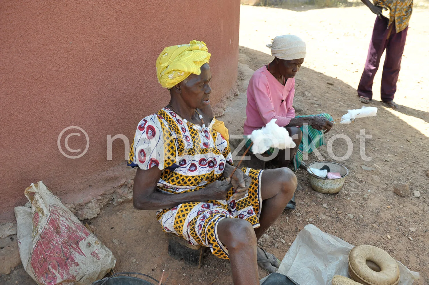 Africa photo — Deux femmes assises contre un mur en terre, l’une en tenue colorée à motifs traditionnels et foulard jaune, l’autre en rose, occupées à