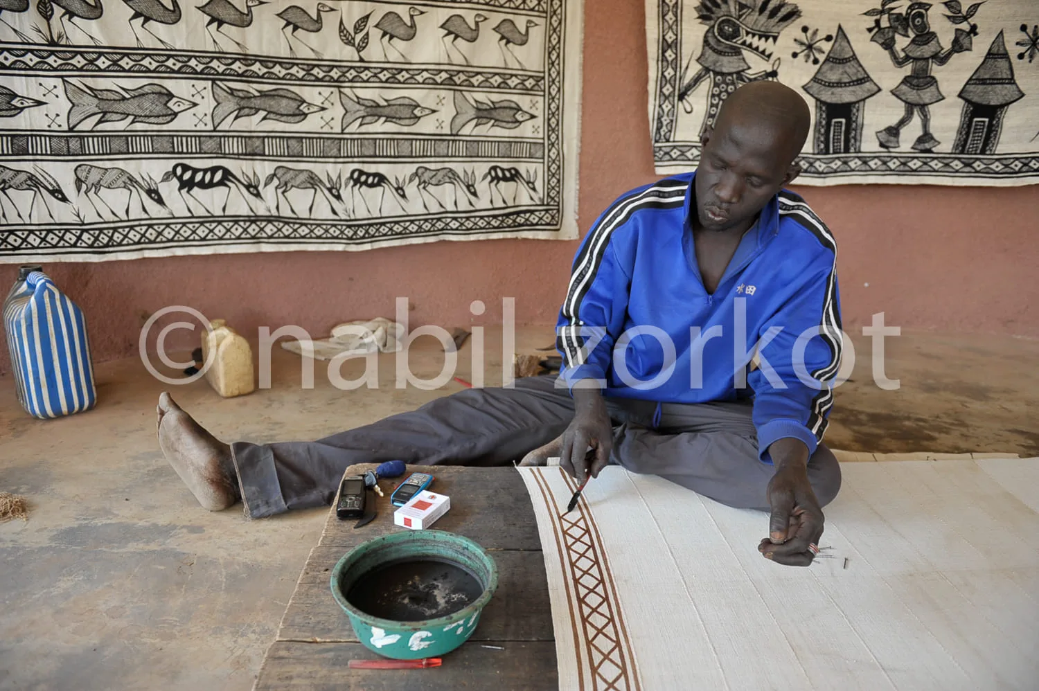 Africa photo — Un artisan assis sur le sol travaille minutieusement sur un tissu blanc étendu, en utilisant des outils de broderie et des pigments nat