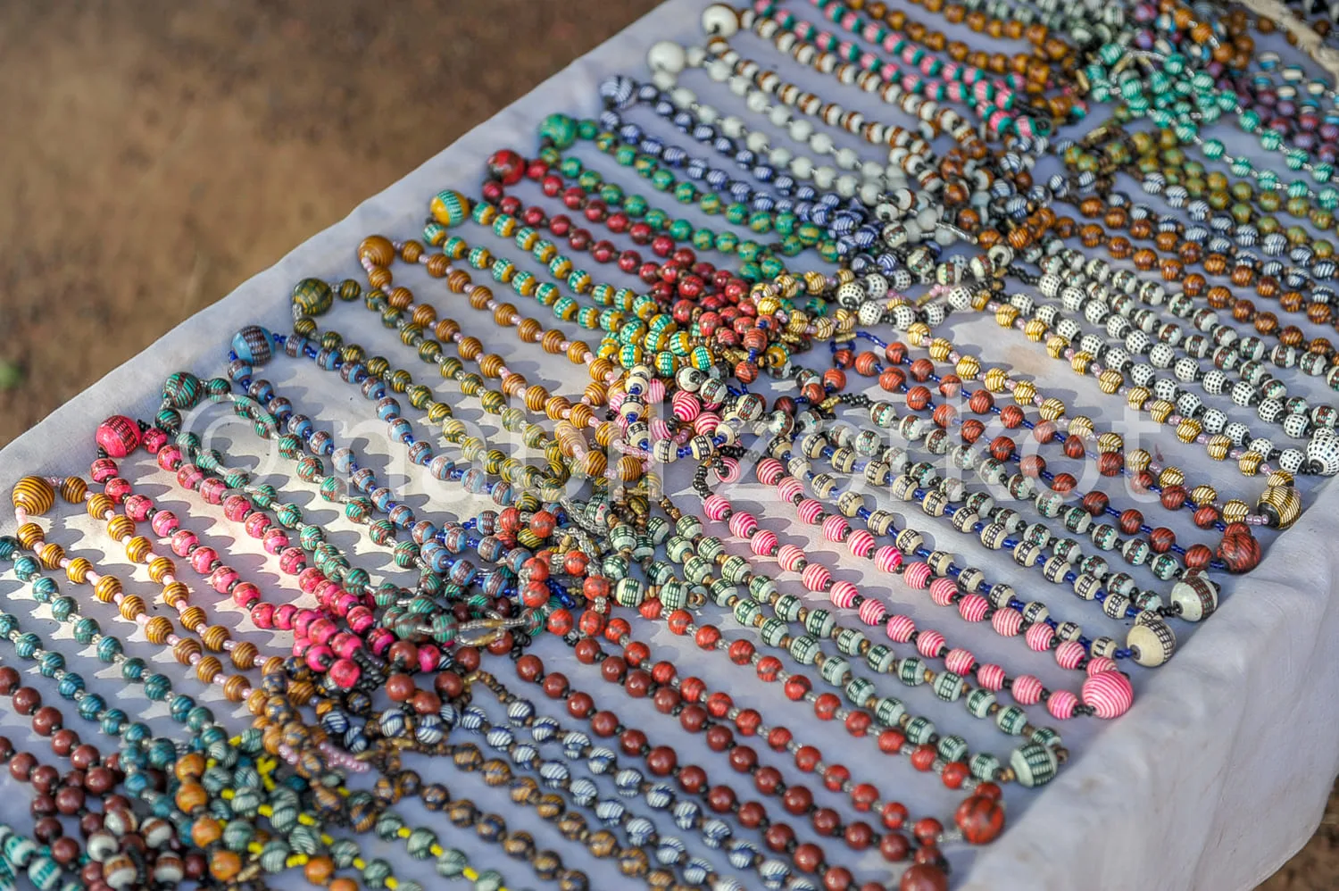 Africa photo — Une étendue de colliers artisanaux composés de perles multicolores aux motifs rayés et unis, étalés sur une surface blanche en extérieu