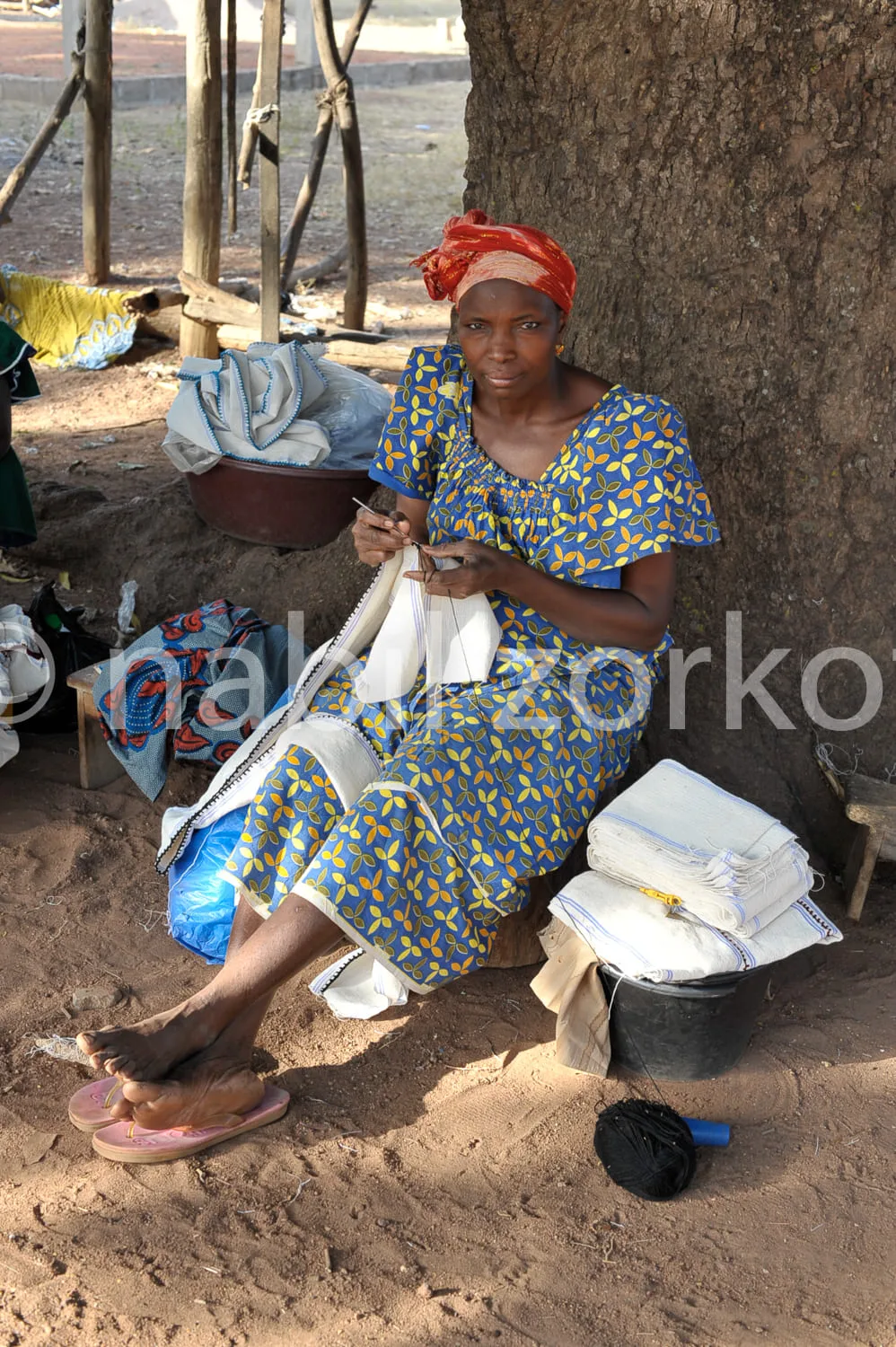 Africa photo — Une femme assise sous un arbre, vêtue d’une robe à motifs bleus et jaunes, porte un foulard rouge. Elle cousant soigneusement un tissu