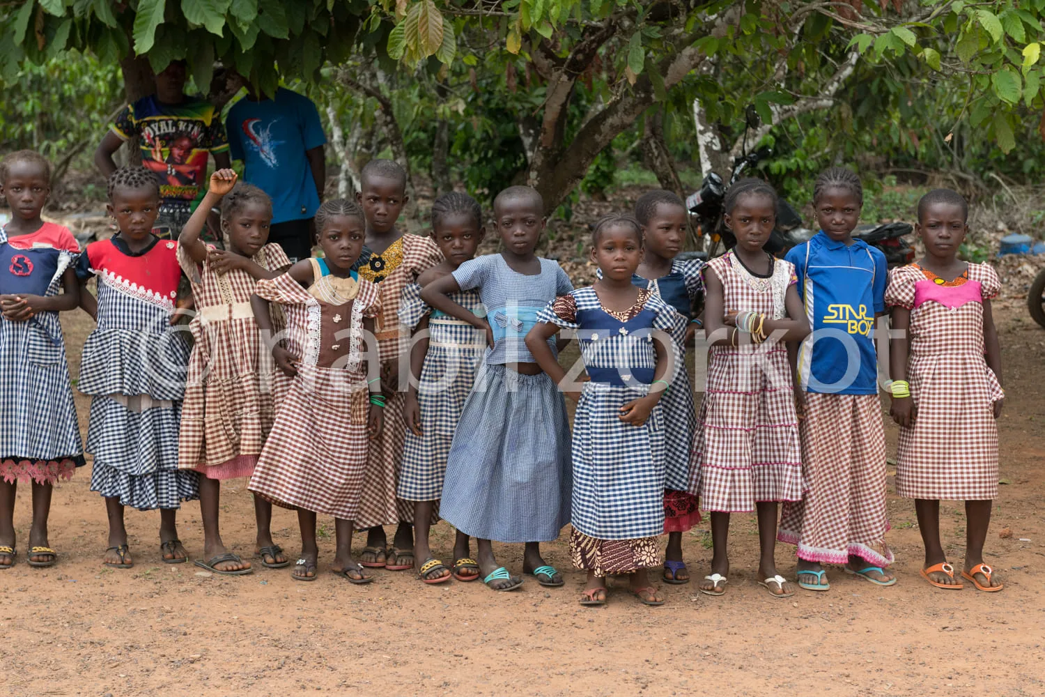 Africa photo — Un groupe de seize enfants, dont huit filles vêtues de robes à carreaux et huit garçons en tenues variées, posent souriants sur un sol