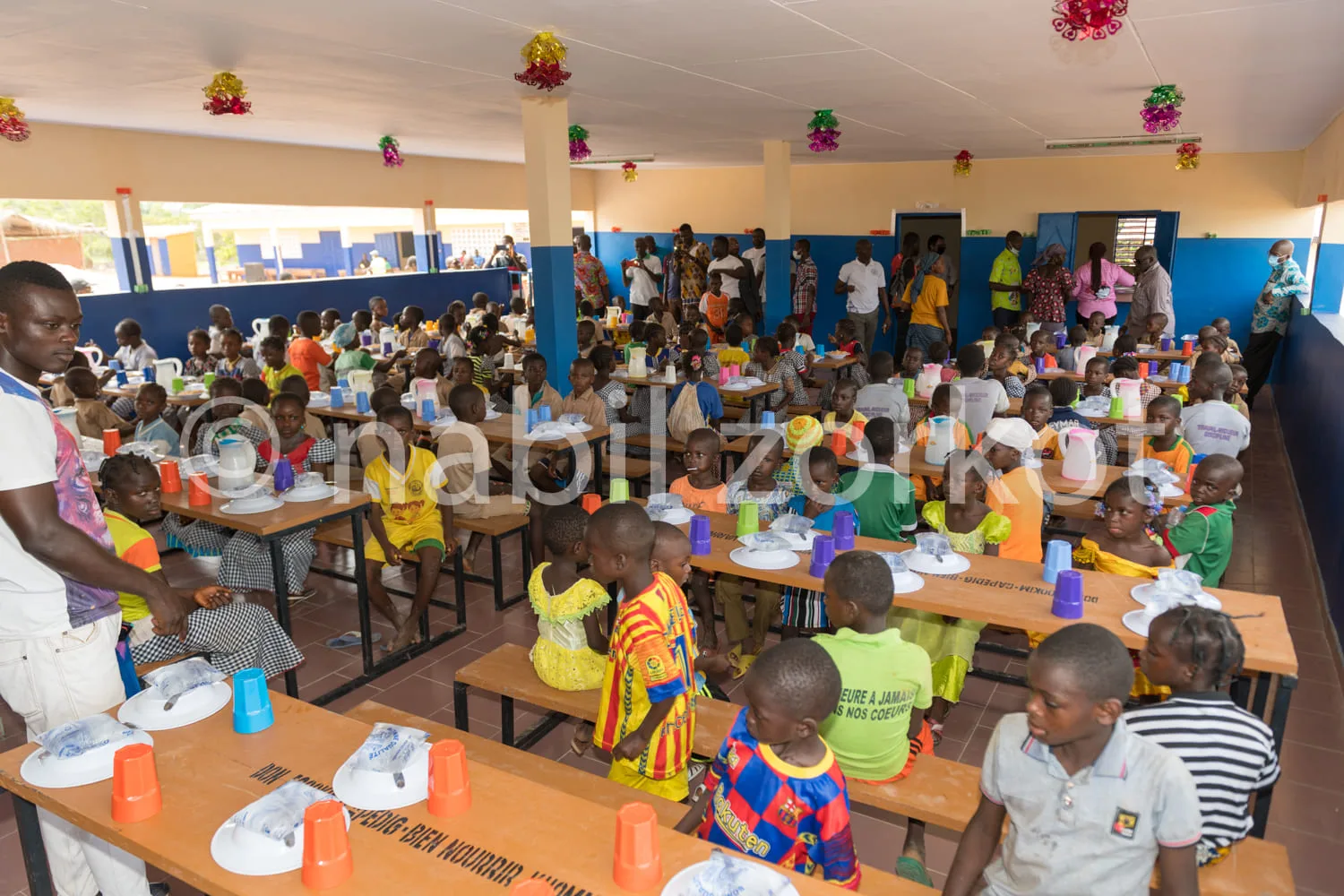 Africa photo — Une grande salle colorée où une centaine d'enfants, assis autour de longues tables, participent à un repas collectif sous la supervisio