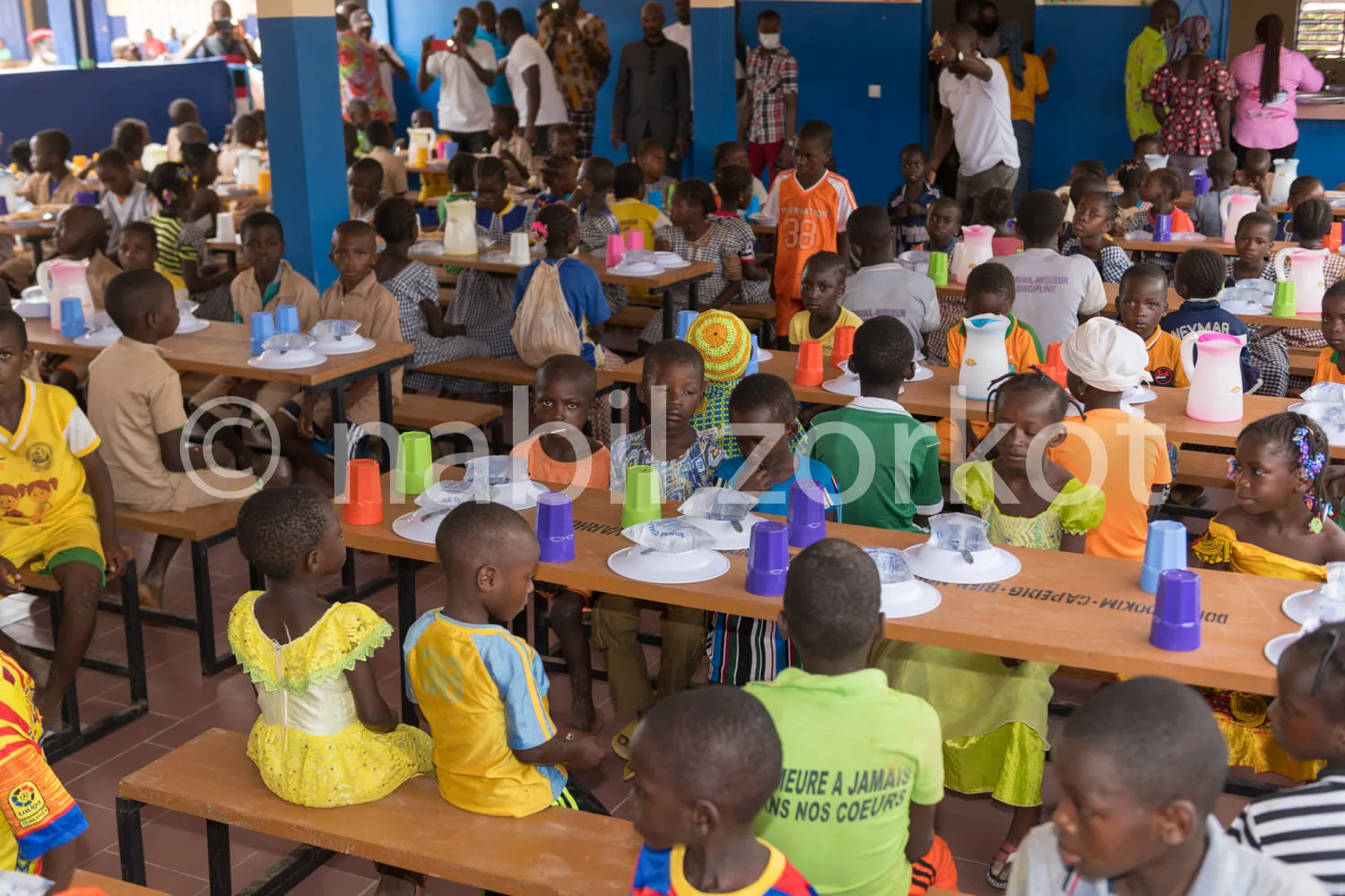 Africa photo — Une grande salle où une centaine d'enfants assises autour de longues tables en bois mangent un repas. Les enfants portent des vêtements