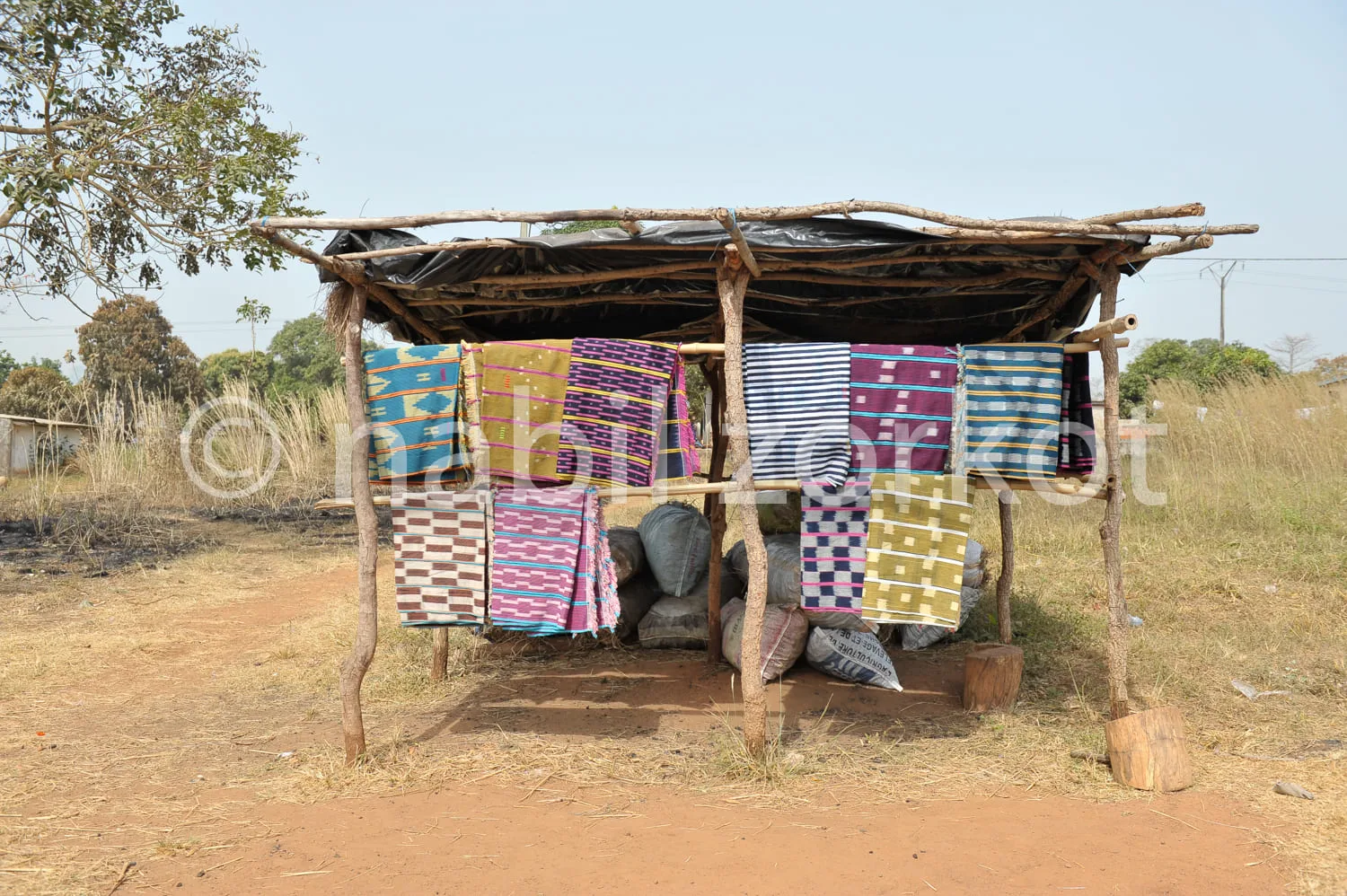 Africa photo — Une petite échoppe en bois recouverte de tôle ondulée, abritant une collection de tissus traditionnels aux motifs variés et colorés acc