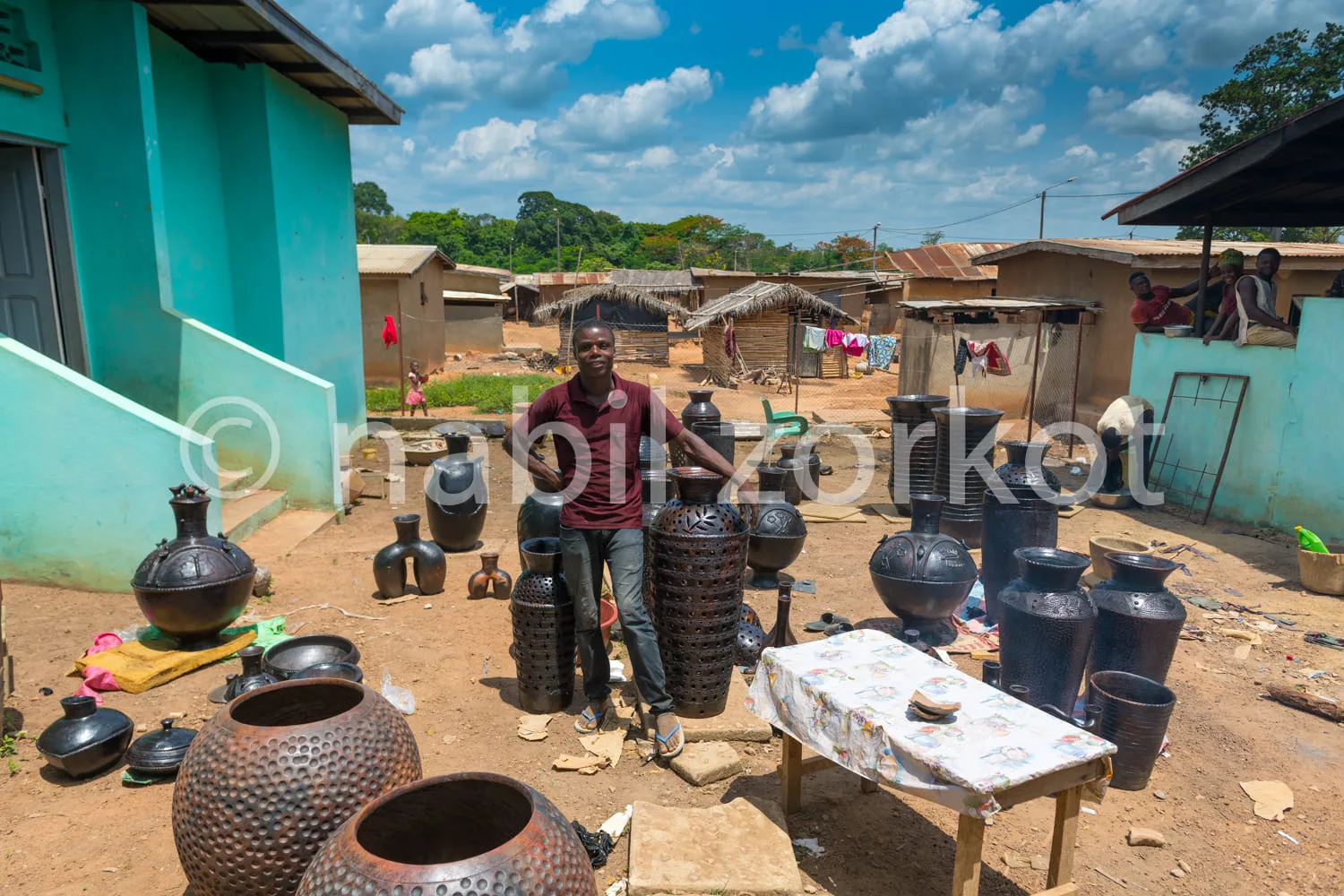 Africa photo — Un artisan debout souriante devant une table et des étagères remplies de pots et jarres en terre cuite de différentes tailles et formes