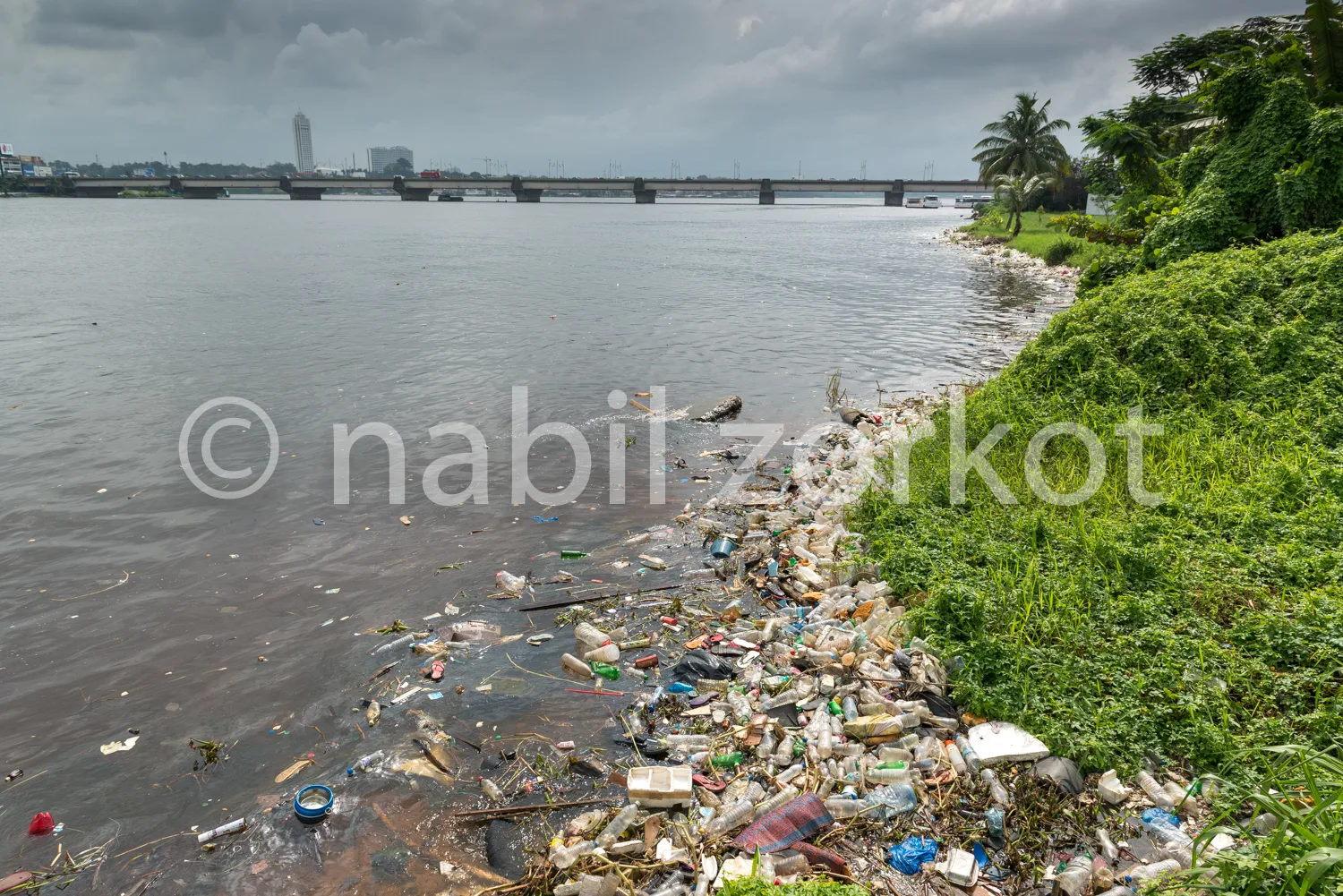 Africa photo — Une berge d'une rivière polluée par des déchets plastiques et divers ordures accumulés près de l'eau, avec un pont et une ville en arri