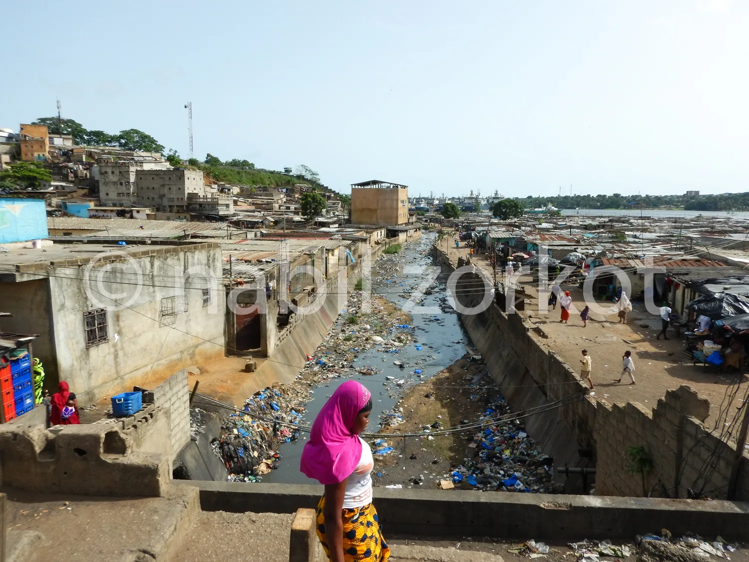 Africa photo — Une femme vêtue d’un hijab rose et d’une robe jaune-multicolore se tient près d’un canal rempli de déchets, entourée de constructions p