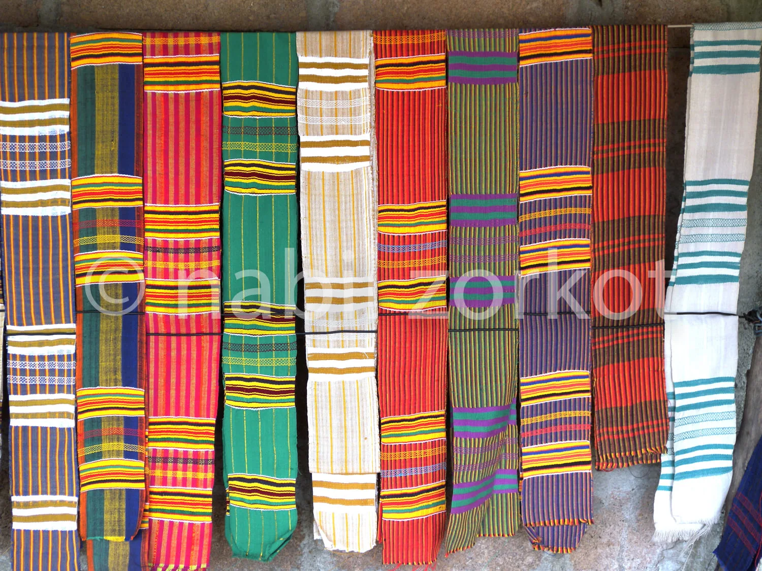 Africa photo — Neuf étoffes kente suspendues en série, chacune présentant des motifs géométriques uniques avec des bandes horizontales et verticales d