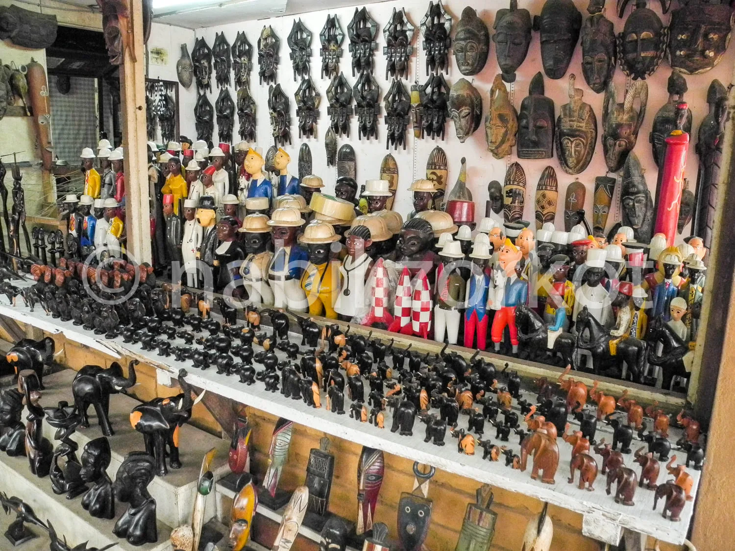 Africa photo — Une étagère remplie de figurines artisanales africaines vêtues de costumes traditionnels aux couleurs vives, entourées de masques en bo