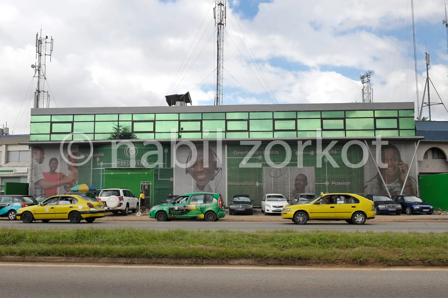 Africa photo — Un bâtiment commercial avec une grande façade verte portant l’enseigne Zorkot en lettres blanches. Trois taxis, dont deux jaunes et un