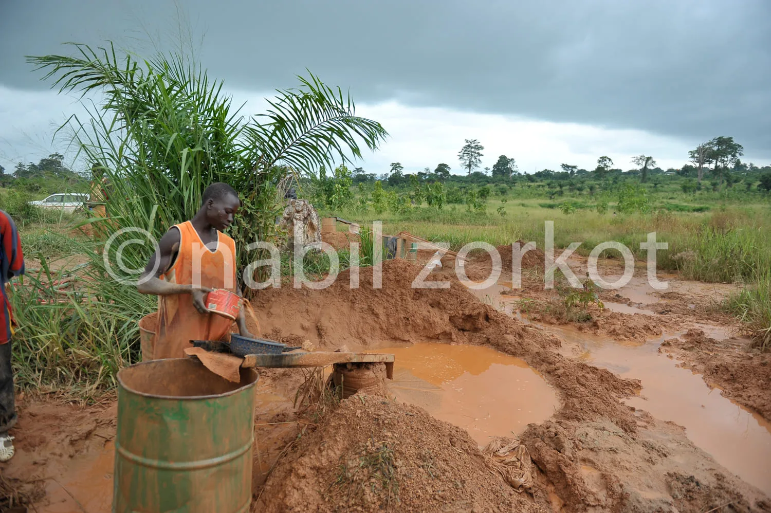 Africa photo — Un homme en tenue orange et marron creuse un trou dans une terre rougeâtre avec une pelle, près d’un seau posé sur le sol. À l’arrière-