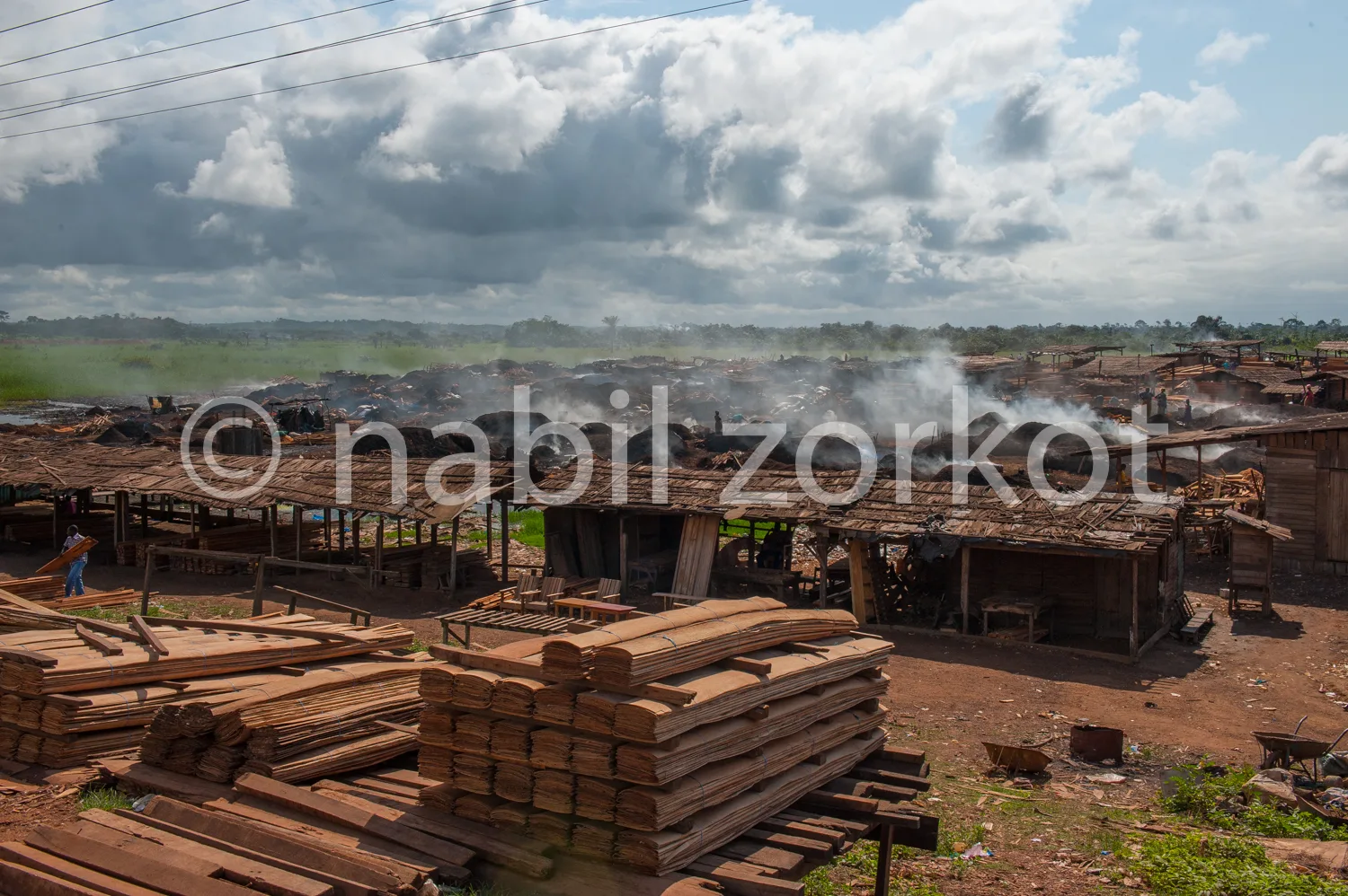 Africa photo — Une vue aérienne d’une scierie artisanale où des fûts de bois sont sciés, produisant de la fumée. Des planches de bois sont empilées au