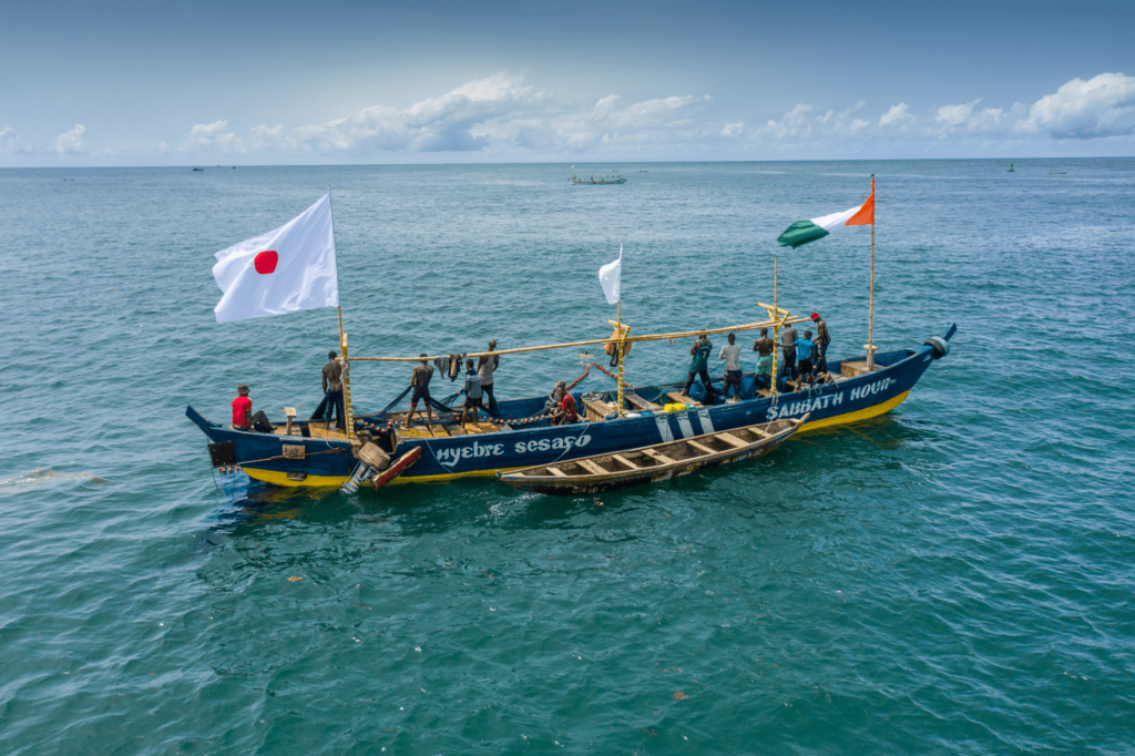 Africa photo — Un bateau traditionnel coloré nommé Nyebre Sesayo navigue en mer avec un équipage de plusieurs personnes. Deux drapeaux flottent : l'un